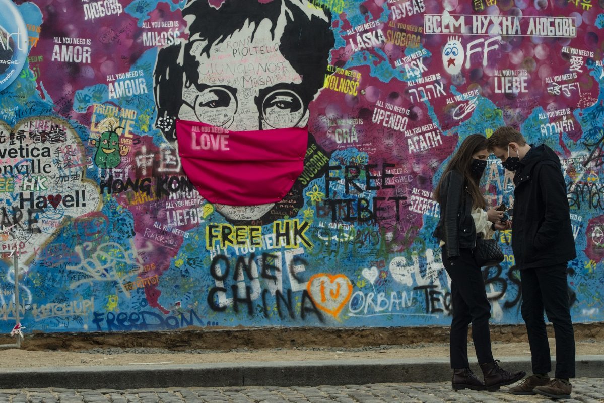 Um casal conversa em frente ao "Muro de Lennon", em Praga, que, em tempos de Covid-19, também usa a sua máscara. Foto Michal Cizek/AFP