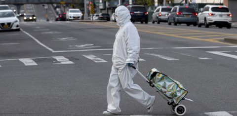Uma mulher vestindo traje e óculos de proteção puxa seu carrinho de compras nas ruas de Queens, em Nova York. Foto Johannes Eisele/AFP