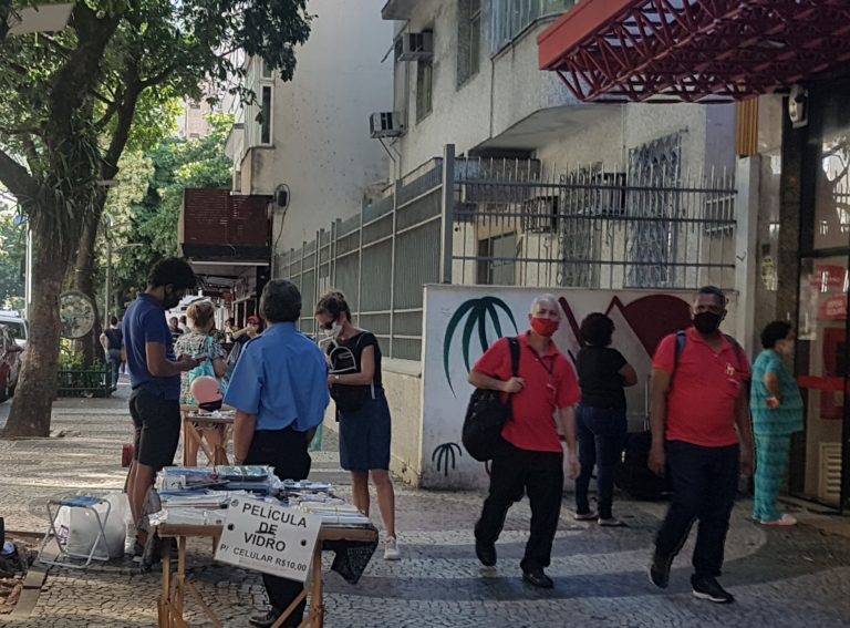 Ambulante mascarado vende máscaras a cliente mascarada, em frente à fila de mascarados em agência bancária na Zona Sul do Rio: isolamento social em queda (Foto: Oscar Valporto)