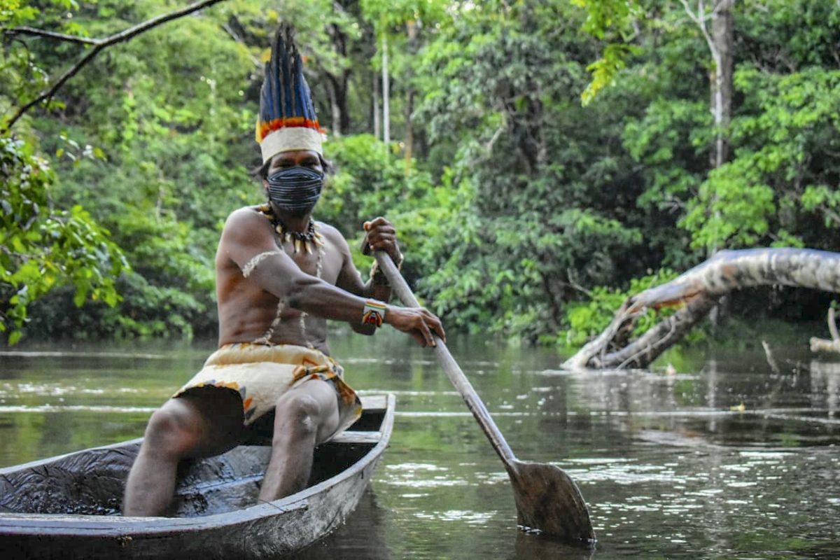 Com sua máscara de proteção,indígena colombiano Huitoto navega em uma balsa ao longo do rio Takana, em Leticia. Foto Tatiana de Nevó/AFP