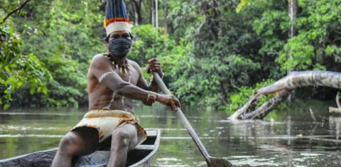 Com sua máscara de proteção,indígena colombiano Huitoto navega em uma balsa ao longo do rio Takana, em Leticia. Foto Tatiana de Nevó/AFP