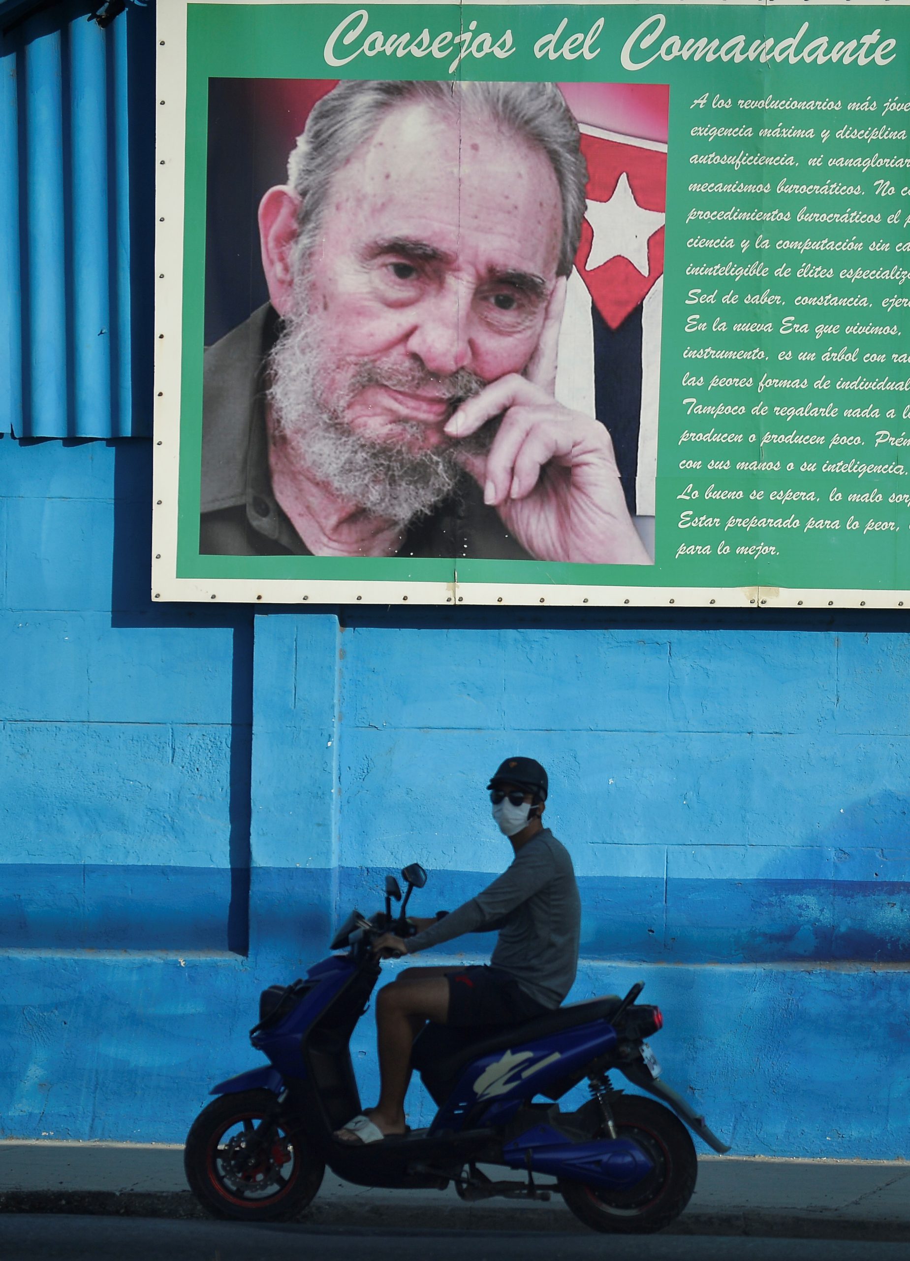 Homem de máscara passa de lambreta, em frente à imagem de Fidel Castro: Havana começa a flexibilizar isolamento. Foto de Yamil Lage (AFP)