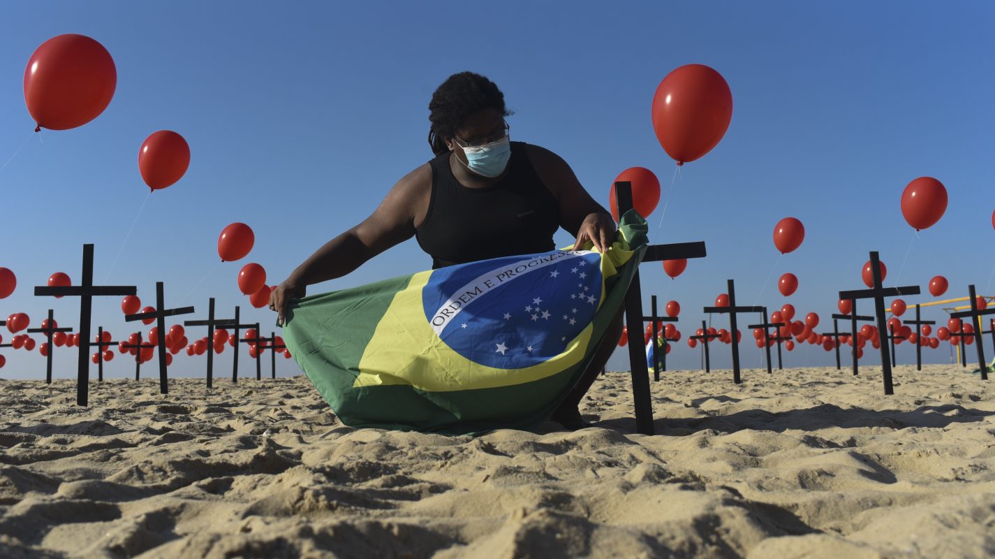 Na praia de Copacabana, no Rio de Janeiro, um homem segura a bandeira brasileira entre balões vermelhos e cruzes em homenagem os cem mil mortos pela covid-19. Foto Fabio Teixeira/Anadolu Agency