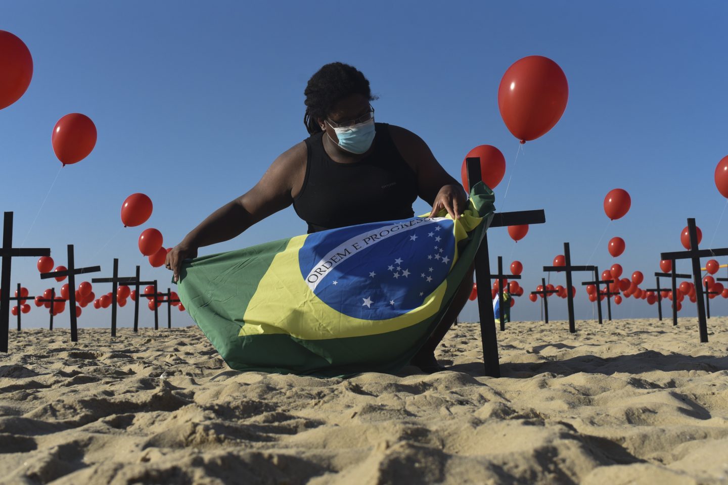 Na praia de Copacabana, no Rio de Janeiro, um homem segura a bandeira brasileira entre balões vermelhos e cruzes em homenagem os cem mil mortos pela covid-19. Foto Fabio Teixeira/Anadolu Agency