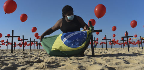 Na praia de Copacabana, no Rio de Janeiro, um homem segura a bandeira brasileira entre balões vermelhos e cruzes em homenagem os cem mil mortos pela covid-19. Foto Fabio Teixeira/Anadolu Agency