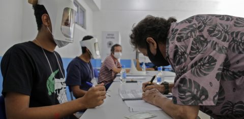 Voluntários usando máscaras e viseiras esperam enquanto um homem assina antes de votar durante o segundo turno das eleições municipais na Rocinha, no Rio de Janeiro. Foto André Coelho/AFP - Novembro de 2020