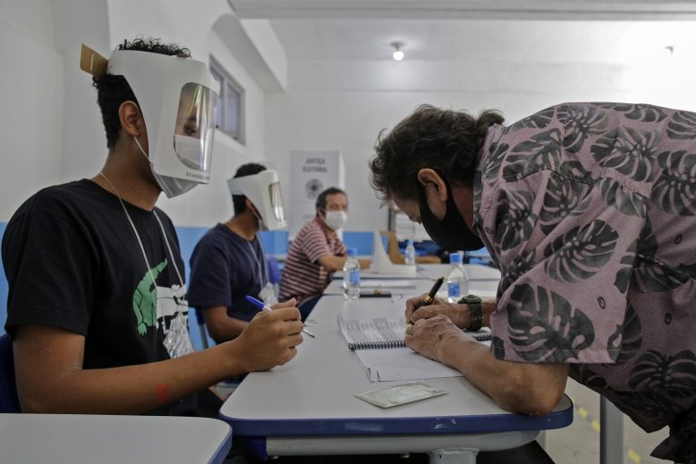 Voluntários usando máscaras e viseiras esperam enquanto um homem assina antes de votar durante o segundo turno das eleições municipais na Rocinha, no Rio de Janeiro. Foto André Coelho/AFP - Novembro de 2020