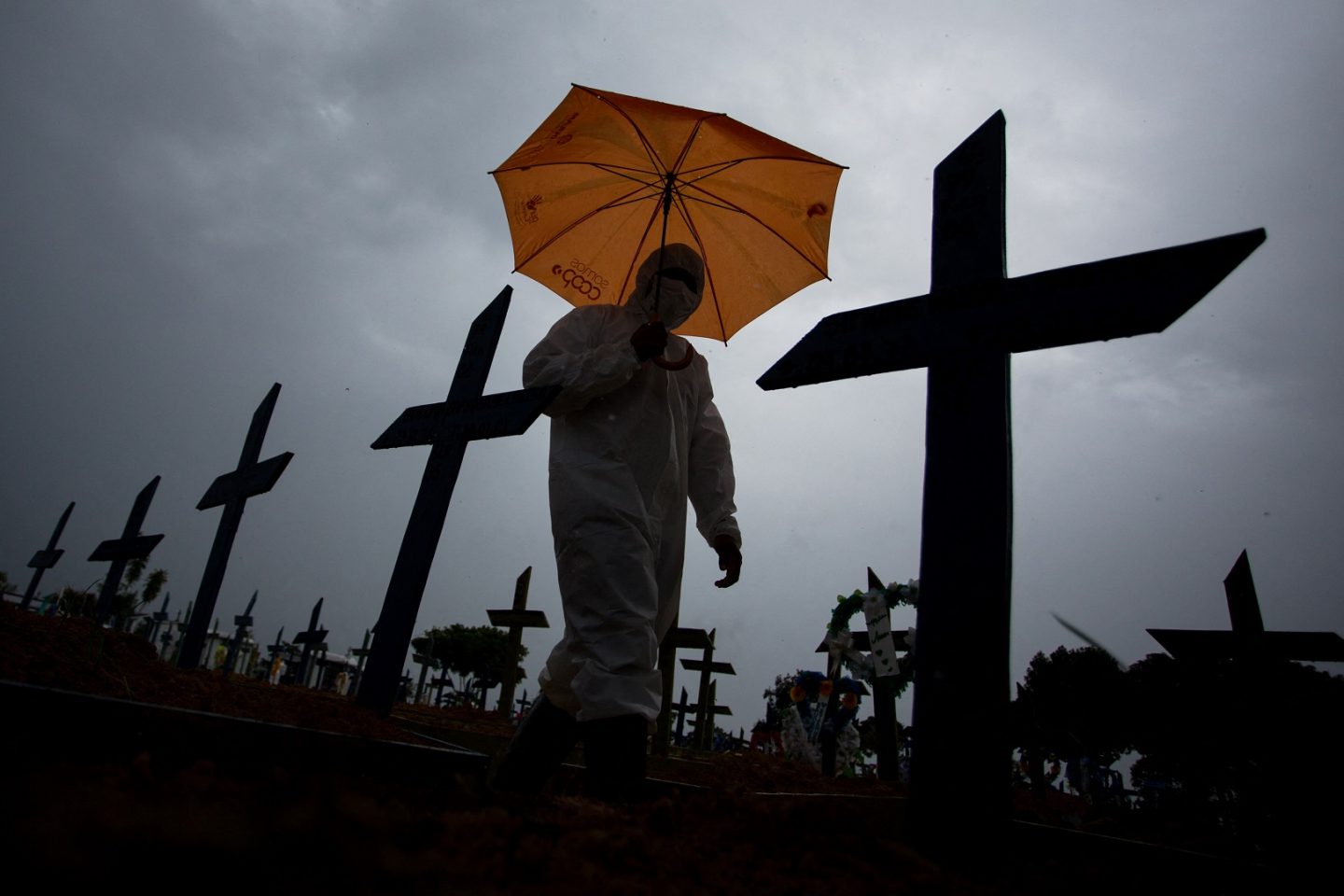 Trabalhador com traje de proteção e guarda-chuva passa por entre os túmulos das vítimas da covid-19 no cemitério de Nossa Senhora Aparecida, em Manaus. Foto Michael Dantas/AFP. Fevereiro/2021