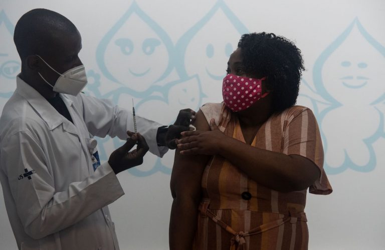 Profissional de saúde, com máscara cirúrgica, vacina moradora do Rio com máscara de pano com camadas: pesquisa com infectados confirmou eficácia da máscara para evitar transmissão (Foto: Fábio Teixeira/NurPhoto/AFP -13/05/2021)