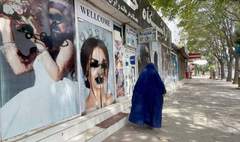 Pessoa vestindo burca passa em frente a cartazes com rostos pichados de mulheres afegãs em um salão de beleza fechado em Cabul