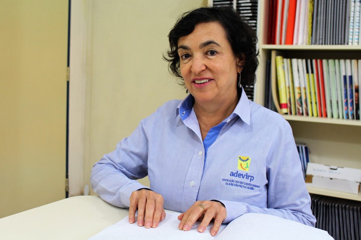 Marlene Cintra é presidente da Associação de Deficientes Visuais de Ribeirão Preto. Está sentada a uma mesa usando uma camisa de manga comprida com a marca da Adevirp. Tem cabelos curtos