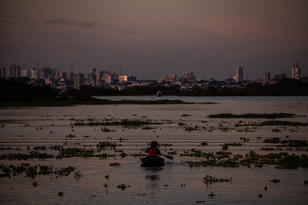 Mulher navega em pequena embarcação pelo Rio Negro, já acima da cota de inundação: cheias no Amazonas ameaçam Manaus (ao fundo) e outras cidades (Foto: Raphael Alves/Amazônia Real)