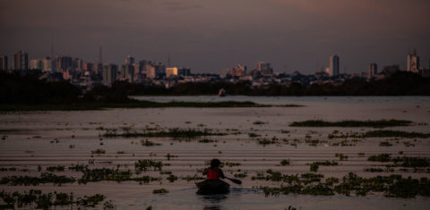 Mulher navega em pequena embarcação pelo Rio Negro, já acima da cota de inundação: cheias no Amazonas ameaçam Manaus (ao fundo) e outras cidades (Foto: Raphael Alves/Amazônia Real)