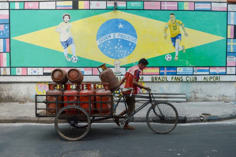 Um trabalhador carrega cilindros de gás em frente a uma arte da bandeira do Brasil desenhada em uma parede em Calcutá, na Índia. Foto Debarchan Chatteriee/NurPhoto, via AFP. Junho/2021