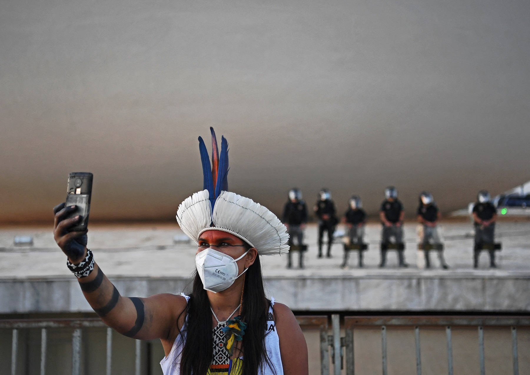 Brasil, América do Sul: mulher indígena, usando máscara contra a covid-19, tira selfie em frente à tropa de choque no prédio do Supremo Tribunal Federal, em Brasília. Foto Carl de Souza/AFP. Agosto/2021