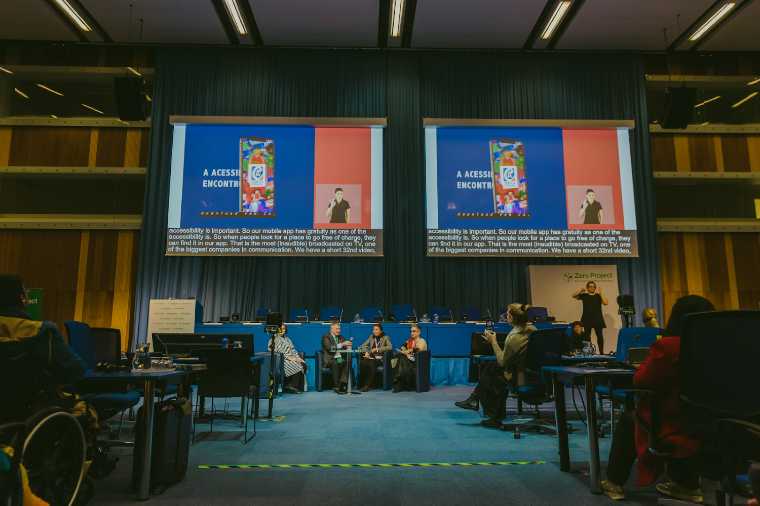 Fotografia colorida de auditório amplo com dois telões exibindo o aplicativo Vem Cá durante Zero Project Conference, em Viena, na Áustria