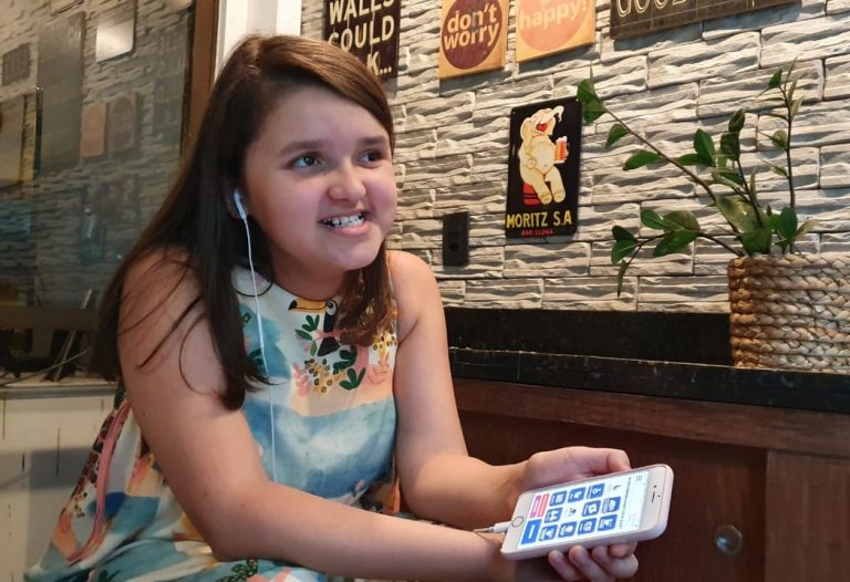 Fotografia da Isabela segurando celular com a mão esquerda, com fones de ouvido conectados para audiodescrição. No celular, a tela mostra a interface do aplicativo Vem Cá. Ela tem expressão sorridente