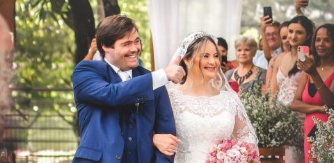 Foto colorida de Dudu do Cavaco e Vitória Rosa em seu casamento. Ambos estão de pé e de braços dados. Ele usa um terno azul e faz um sinal de positivo com a mão direita em direção ao lado direito da foto. Ela está usando um vestido de noiva branco e segura um buquê de flores rosas com a mão esquerda. Ambos sorriem e olham para a direita