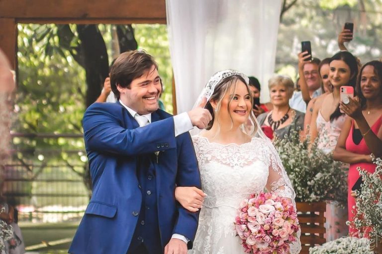 Foto colorida de Dudu do Cavaco e Vitória Rosa em seu casamento. Ambos estão de pé e de braços dados. Ele usa um terno azul e faz um sinal de positivo com a mão direita em direção ao lado direito da foto. Ela está usando um vestido de noiva branco e segura um buquê de flores rosas com a mão esquerda. Ambos sorriem e olham para a direita