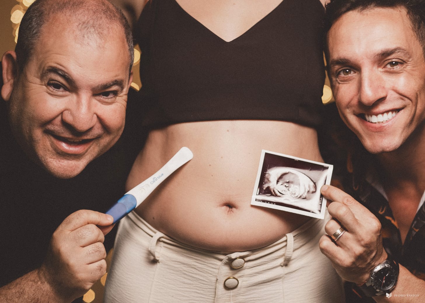Foto colorida de Jarbas e Mikael segurando foto do ultrassom e de teste de gravidez próxima a barriga de Jéssica. Ambos olham para a foto e sorriem
