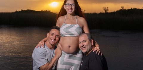 Foto colorida de Jarbas e Mikael com Jéssica. Ambos estão abaixados com os rostos próximos da barriga de Jéssica, enquanto sorriem. Ao fundo, pôr do Sol e lago.