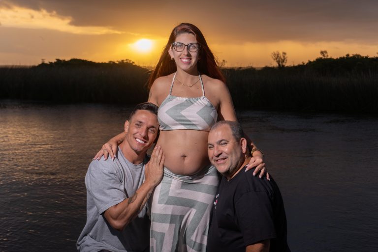 Foto colorida de Jarbas e Mikael com Jéssica. Ambos estão abaixados com os rostos próximos da barriga de Jéssica, enquanto sorriem. Ao fundo, pôr do Sol e lago.