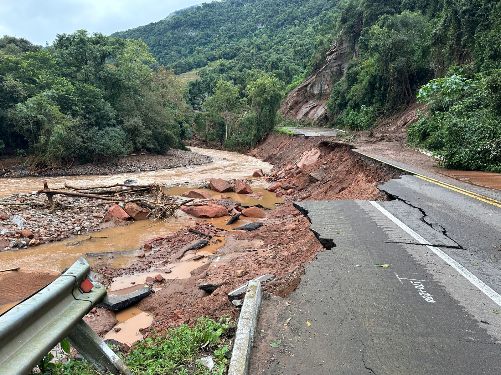 Foto colorida da pista da BR-116, no município de Caxias do Sul, destruída pela enchente. A imagem mostra a pista destruída por deslizamento de terra, com lama e poças de água no lado esquerdo da imagem. Ao fundo, árvores e serra