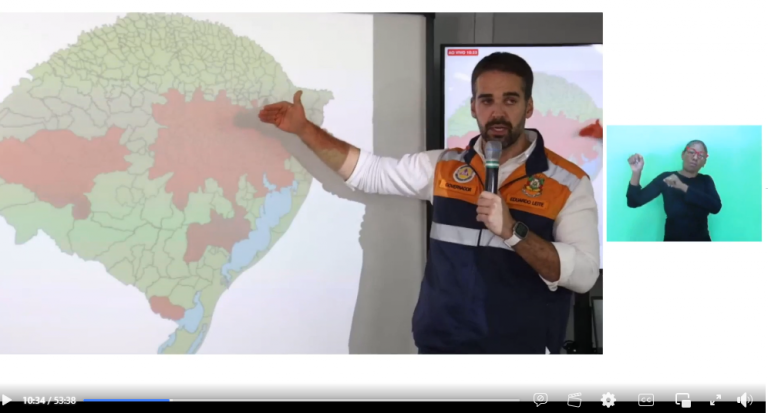 Captura de tela colorida do governador do Rio Grande do Sul Eduardo Leite. Leite está de pé ao lado de um mapa do RS com partes em vermelho nas regiões central e metropolitana de Porto Alegre. Ele segura um microfone e aponta com a mão direita para o mapa. Ele usa um colete da defesa civil e uma camiseta branca por baixo. Leite é um homem branco com barba e cabelos pretos. Ao seu lado, em uma tela pequena aparece uma intérprete. Ela é uma mulher negra com uma camiseta preta e está sinalizando em Libras, em um fundo verde-azulado.