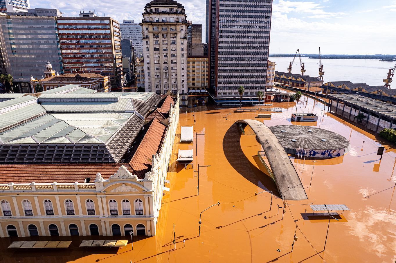 Foto colorida do Centro Histórico de Porto Alegre invadido pelas águas. Na imagem, os prédios estão com água cobrindo suas entradas. A cor de água é marrom e muitas estruturas no canto direito estão submersas. No canto superior direito, ao fundo é possível ver o Guaíba.