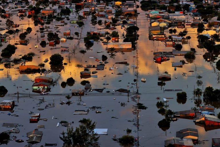 Foto colorida da região Metropolitana de Porto Alegre submersa após enchente. A água toma a maior parte da imagem, com apenas alguns pontos em que aparecem árvores e casas não inundadas.