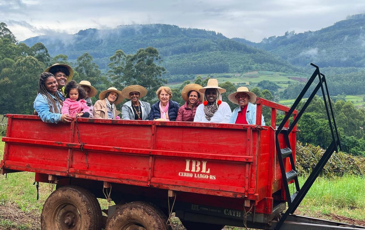 Foto colorida de pessoas em cima de um carretel vermelho. Elas usam chapéu e sorriem para a câmera. Ao fundo, sítio com paisagens e montanhas