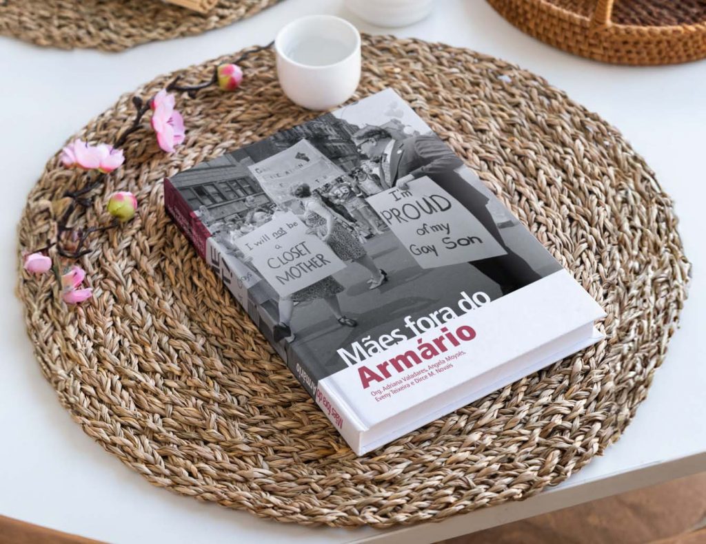 Foto colorida de livro Mães fora do Armário. O livro está sobre uma mesa. A capa dele mostra pessoas em uma manifestação em fotografia preto e branco e o título, com letras em branco e a palavra "Armário" em vermelho