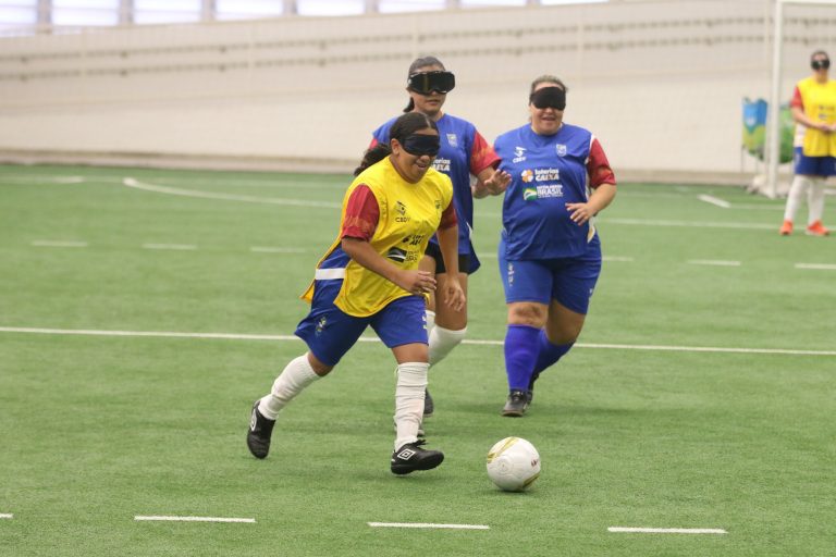 Foto colorida de atletas disputando jogo de futebol de cegas em São Paulo. No centro da imagem, uma mulher cega de camiseta amarela corre com a bola, enquanto sorri. Ela usa bermuda azul e meias brancas, além de proteção nos olhos. Ao fundo, aparecem duas mulheres vestidas de camiseta, bermuda e meias azuis, elas correm em direção à bola.