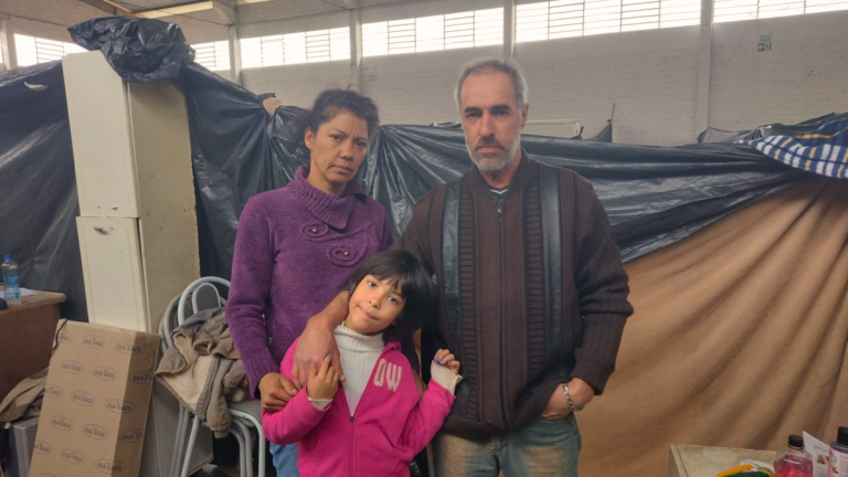 Foto colorida de Adelar, Valeria e Pamela em abrigo de Lajeado. Eles estão abraçados e usam roupa de frio, ao fundo barraca de abrigo.