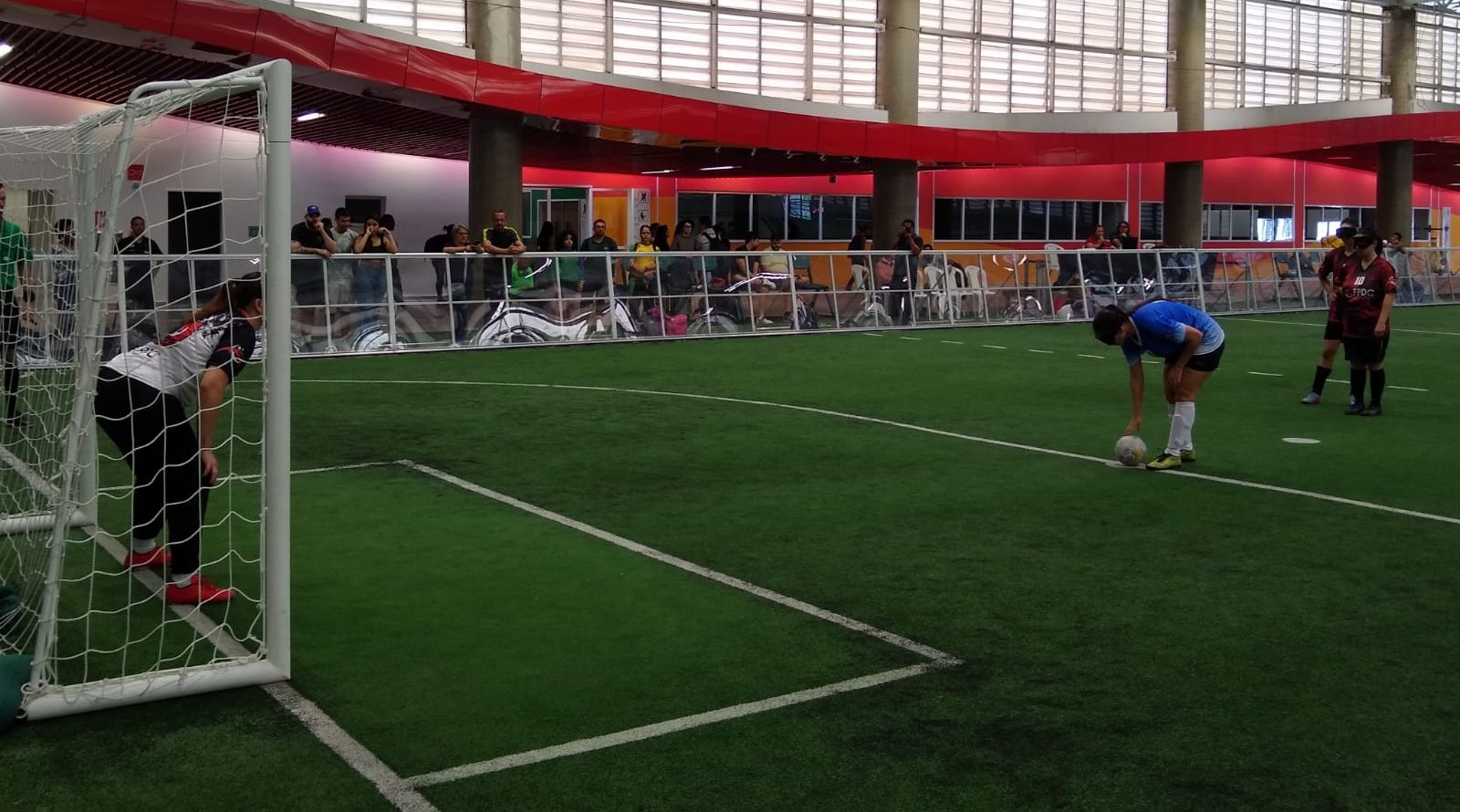 Foto colorida de Geisa Farini se preparando para cobrança de pênalti em partida de futebol de cegas. No canto direito, Geisa de camiseta azul prepara a bola na marca do pênalti, no canto esquerda, goleira se prepara para tentar defender a bola