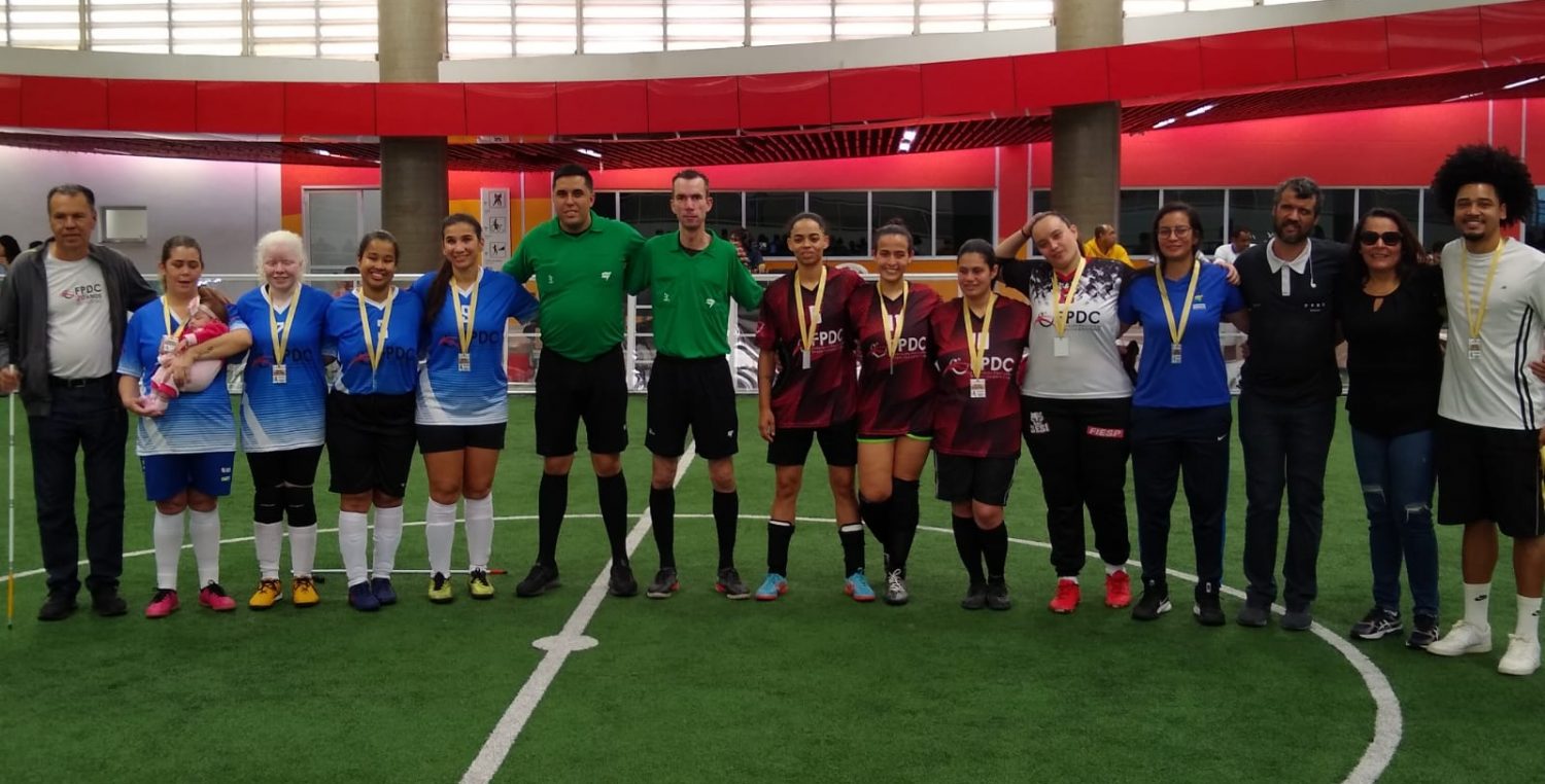 Foto colorida de atletas que participaram de partida de futebol de cegas amistosa. Perfilamento final com medalhas entregues. Da esquerda para a direita, Sr. Benedito Franco, jogadoras time azul, dois árbitros da partida, time vermelho, goleiros, equipe diretora Federação, técnicos