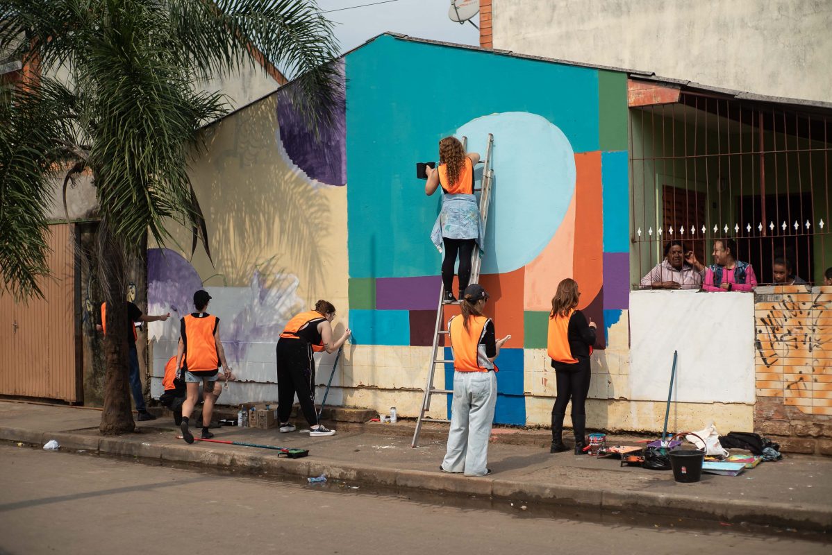 Foto colorida de voluntários do Paredes com Propósito. Na imagem, diversos voluntários pintam parede de uma casa. Eles usam coletes laranjas. Ao lado, aparecem moradores observando o trabalho