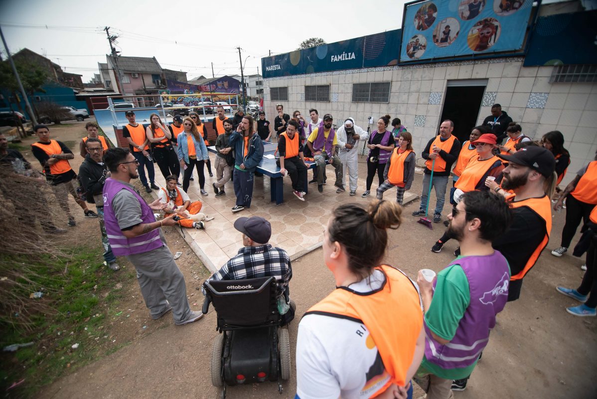 Foto colorida de reunião entre voluntários e membros da Vila dos Papeleiros. Na imagem, diversas pessoas aparecem em pé