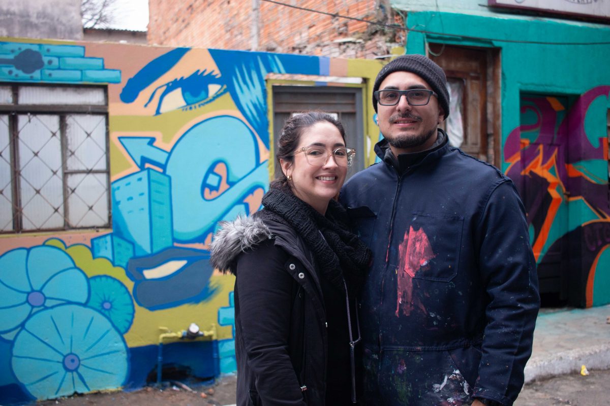 Foto colorida de Jotapê e Kami em frente a casa pintada pelo projeto. Jotapê é um homem branco e usa óculos, touca e um agasalho. Kami é uma mulher branca com cabelo castanho e usa um casaco. Ao fundo, parede pintada colorida