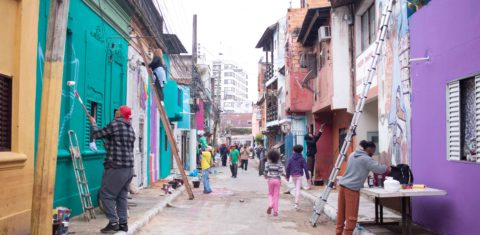 Foto colorida de mutirão do Paredes com Proposito. Na imagem, diversas pessoas aparecem pintando residências em rua do Quilombo do Areal, em Porto Alegre. Artistas pintam paredes, enquanto moradores passeiam pelo local