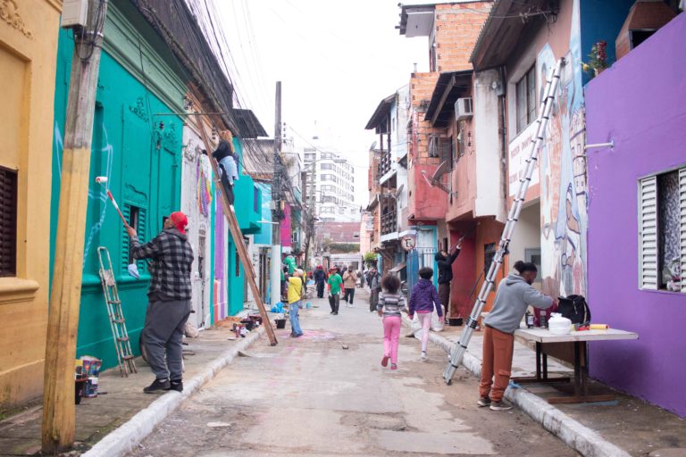 Foto colorida de mutirão do Paredes com Proposito. Na imagem, diversas pessoas aparecem pintando residências em rua do Quilombo do Areal, em Porto Alegre. Artistas pintam paredes, enquanto moradores passeiam pelo local