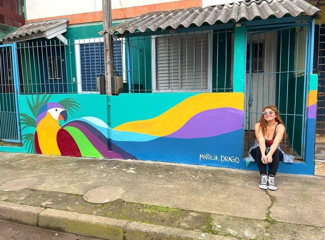 Foto colorida de fachada de casa pintada pelo projeto Paredes com Propósito. No canto inferior direito, Marília Drago aparece sentada. Ela é uma mulher branca com cabelos ruivos e usa uma camiseta laranja, ela olha para a câmera e sorri