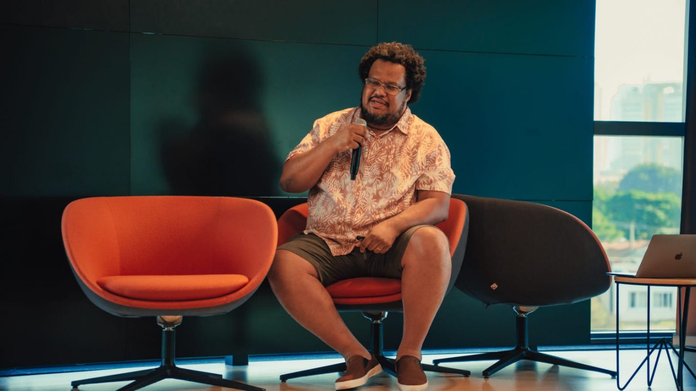 Foto colorida de Fábio Souza, conhecido como Tio Faso. Ele é um homem negro de cabelo afro e está sentado em uma cadeira com um microfone na mão. Ele usa uma camisa listrada em branco e laranja e uma bermuda verde musgo. Ao fundo, parede e ao lado dele outras cadeiras.