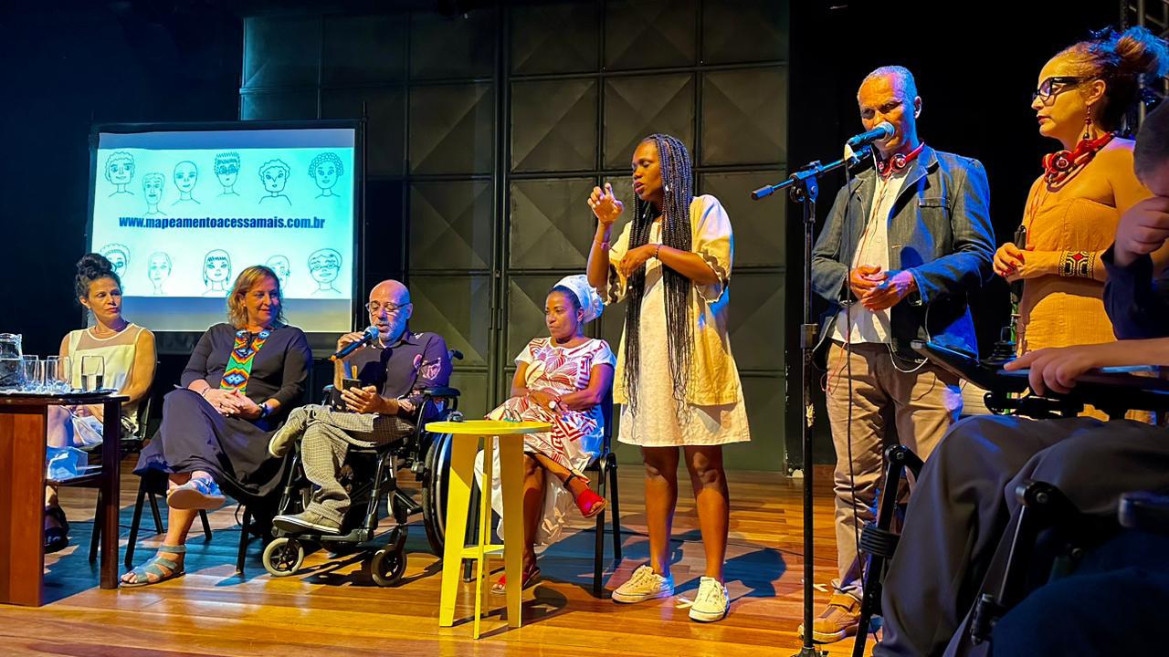 Foto colorida do evento de lançamento do mapeamento Acessa Mais. Na imagem, Edu O. aparece sentado e com o microfone na mão, no seu lado direito duas mulheres aparecem sentadas, e uma do lado esquerdo. No canto direito da imagem, uma mulher negra aparece em pé sinalizando em Libras. Do lado da intérprete, outros dois homens aparecem em pé. Ao fundo, data show mostra apresentação do mapeamento