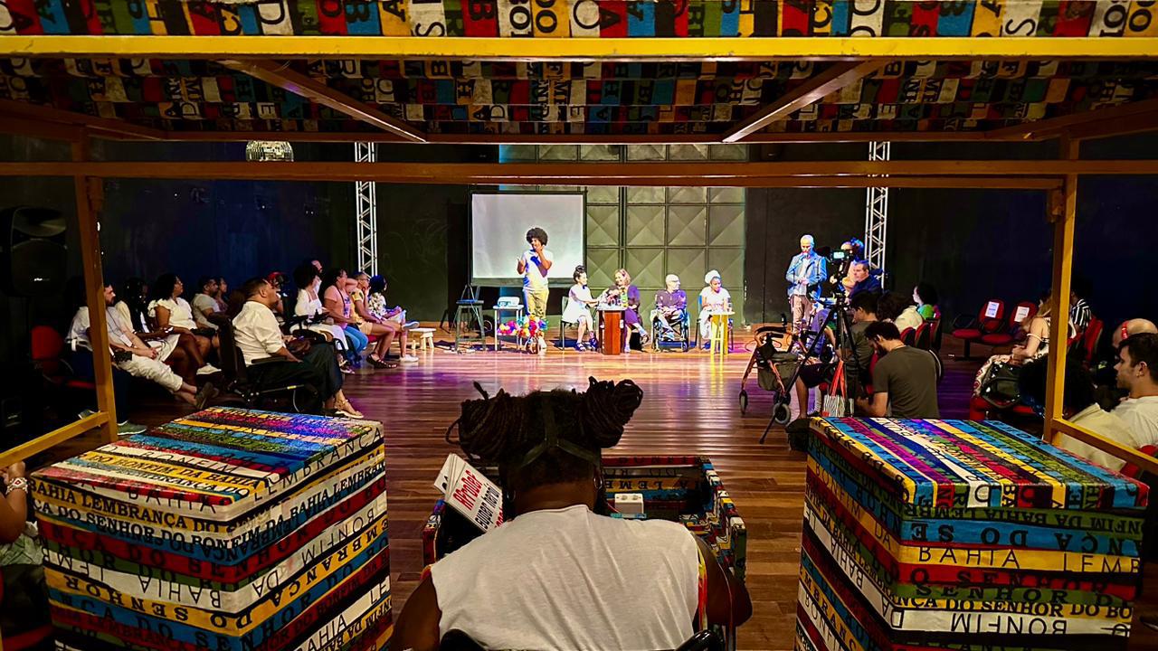 Foto colorida de Teatro da UFBA. No centro da imagem, artistas def apresentam o projeto do mapeamento. No primeiro plano, uma pessoa controla o sistema de som e iluminação. No canto esquerdo e direito, pessoas aparecem sentadas e assistem a apresentação