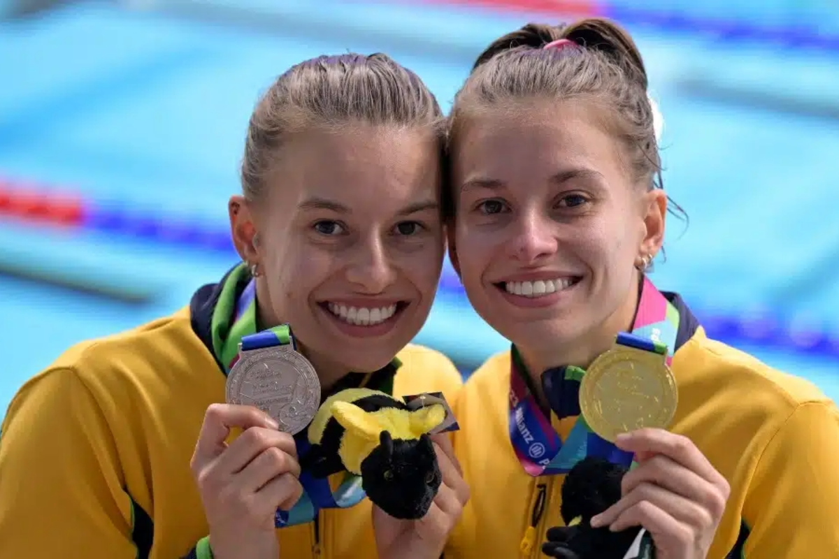 Foto colorida das nadadoras Beatriz e Débora, diagnosticadas com deficiência intectual na infância. Elas são mulheres brancas, com cabelo castanho e seguram suas medalhas do Mundial de Natação Paralímpica. Elas olham para a câmera e sorriem. Ao fundo piscina de natação