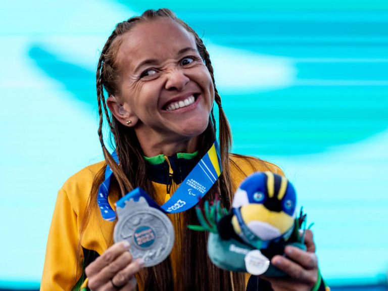 Foto colorida da atleta paralímpica Samira Brito. Ela é uma mulher negra com cabelo dreads. Ela segura sua medalha de prata no Parapan de Santiago 2023, enquanto sorri