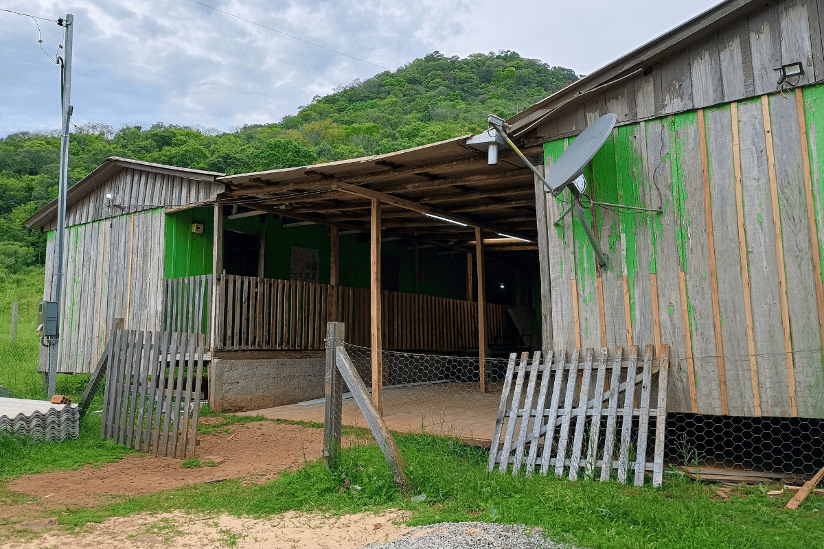 Foto colorida da fachada da escola Augusto Ope da Silva, em Santa Maria. A escola é de madeira, com duas construções interligadas por um telhado coberto. As paredes estão desbotadas e apresentam tons de verde. Ao fundo, encosta de morro.