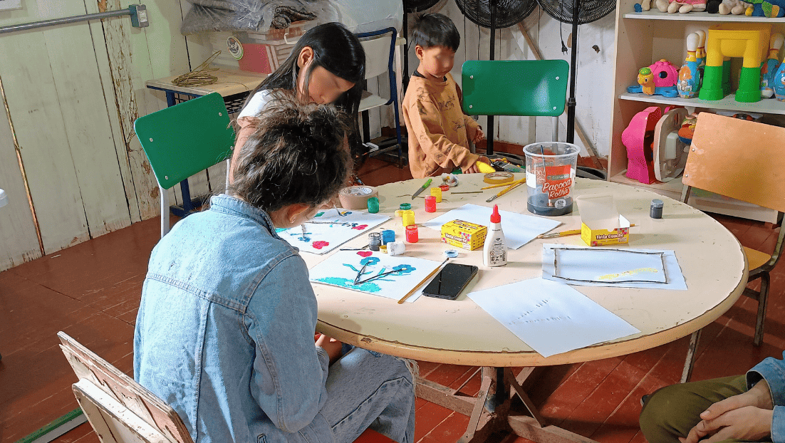 Foto colorida de crianças fazendo pinturas em oficinas promovida por estudantes da UFSM. Na imagem, aparecem duas crianças indígenas e uma estudante sentada de costas