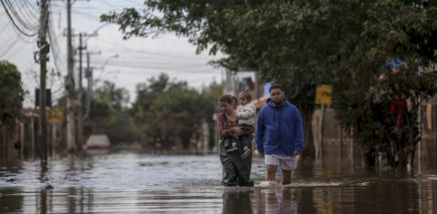 Foto colorida de mulher com criança no colo andando por rua alagada em Porto Alegre. Ao lado delas, aparece um homem também andando em meio às águas. Desastre mostram relação entre saúde materna e mudanças climáticas